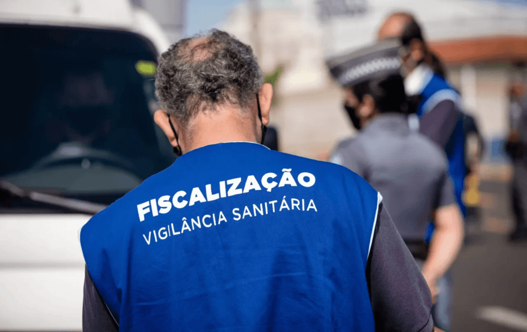 Agente da Vigilância Sanitária durante uma fiscalização, vestindo um colete azul com a inscrição "Fiscalização Vigilância Sanitária", destacando a importância do controle de pragas como a desratização.